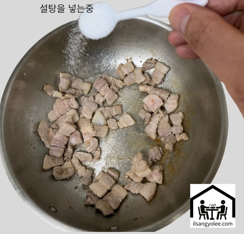 노릇하게 볶이는 돼지고기 위에 흰색 숟가락을 사용하여 설탕을 뿌리고 있는 모습.